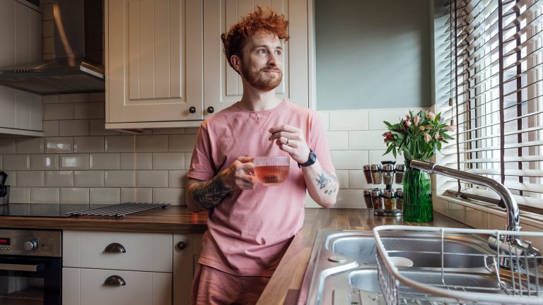 Eine Person steht in der Küche. In der Hand hält sie ein Teeglas, in dem gerade ein Tee zieht.