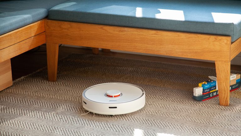 Ein Roborock S5 Max f&auml;hrt &uuml;ber einen Teppich vor einer Sitzbank.