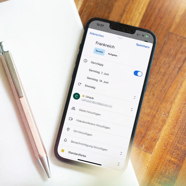 Ein iPhone liegt auf einem Notizblock. Auf dem Display ist ein erstellter Eintrag im Google Kalender f&uuml;r einen Urlaub zu sehen.