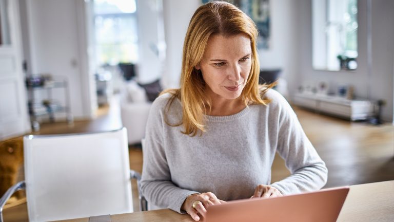 Eine Person sitzt an einem Esstisch im Wohnzimmer vor einem Laptop.