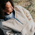Eine Person liegt mit geschlossenen Augen in einem Fleecepullover in einer Sandd&uuml;ne mit einigen Gr&auml;sern.