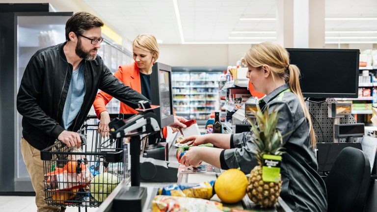 Eine Person steht mit einem Einkaufswagen an der Supermarktkasse, Waren liegen bereits auf dem Band. Die Person legt weitere Waren darauf.