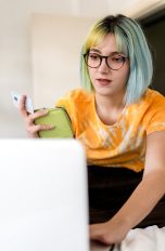 Eine Person sitzt auf einem Bett vor dem geöffneten Laptop. In der Hand hält sie ein Portemonnaie und eine Kreditkarte.
