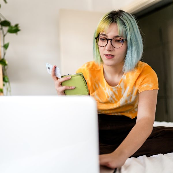 Eine Person sitzt auf einem Bett vor dem ge&ouml;ffneten Laptop. In der Hand h&auml;lt sie ein Portemonnaie und eine Kreditkarte.
