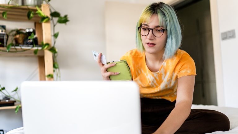 Eine Person sitzt auf einem Bett vor dem ge&ouml;ffneten Laptop. In der Hand h&auml;lt sie ein Portemonnaie und eine Kreditkarte.