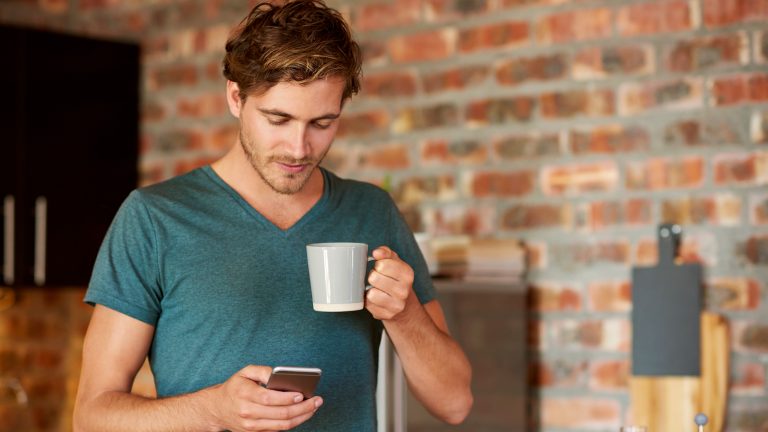 Eine Person steht in einer K&uuml;che, h&auml;lt eine Kaffeetasse in der Hand und schaut auf das Smartphone in ihrer Hand.