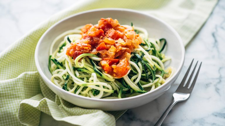 Auf einem Teller befinden sich Zucchini-Nudeln, die teilweise von etwas Tomatensauce bedeckt werden.