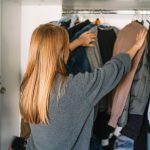 Eine Person steht vor einem Kleiderschrank und schiebt mit beiden H&auml;nden Pullover auf B&uuml;geln darin auseinander.