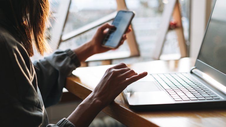 R&uuml;ckansicht einer Person, die an einem Schreibtisch vor einem Laptop sitzt. In der Hand h&auml;lt sie auch ein Smartphone.