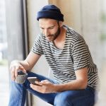 Eine Person sitzt an einem Fenster, h&auml;lt eine Tasse in der Hand und schaut auf das Smartphone, welches sie in der anderen Hand h&auml;lt.