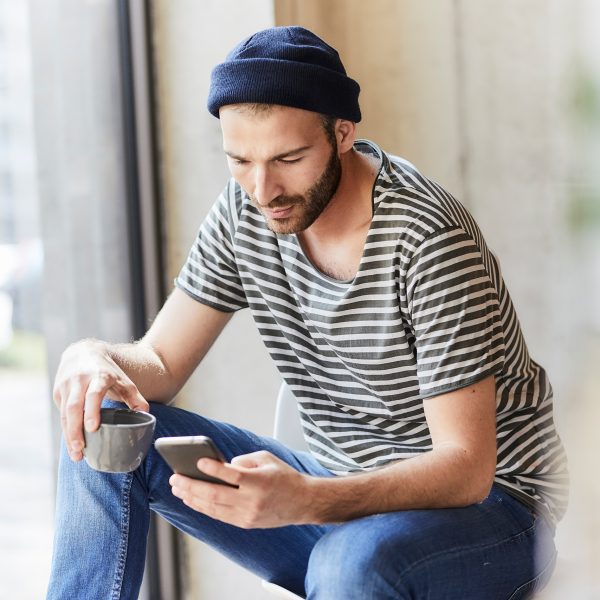Eine Person sitzt an einem Fenster, h&auml;lt eine Tasse in der Hand und schaut auf das Smartphone, welches sie in der anderen Hand h&auml;lt.