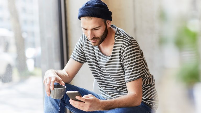 Eine Person sitzt an einem Fenster, h&auml;lt eine Tasse in der Hand und schaut auf das Smartphone, welches sie in der anderen Hand h&auml;lt.