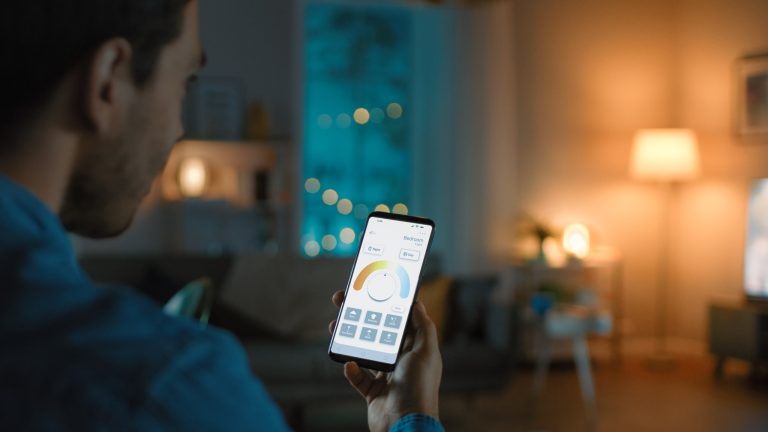 Eine Person sitzt in einem Haus mit atmosph&auml;rischer Beleuchtung. In der Hand h&auml;lt sie ein Smartphone, auf dem eine App zur Steuerung des Smart Homes ge&ouml;ffnet ist.