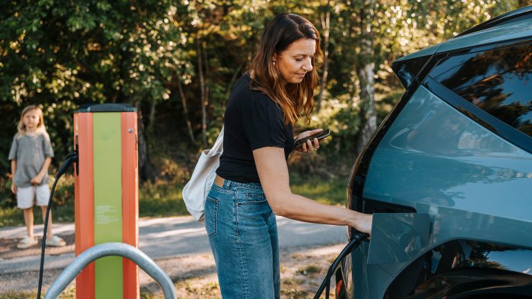 Eine Person steht an einer Lades&auml;ule und steckt einen Stecker in ein E-Auto, um es zu laden.