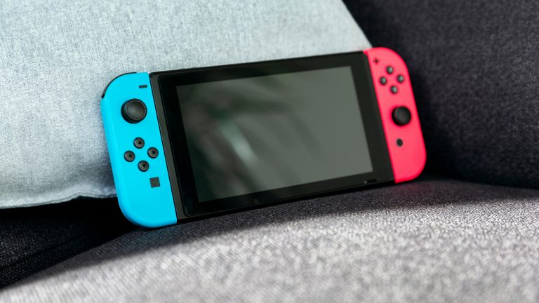 Eine Nintendo Switch mit blauen und roten JoyCons lehnt an einem Kissen auf einer Couch.