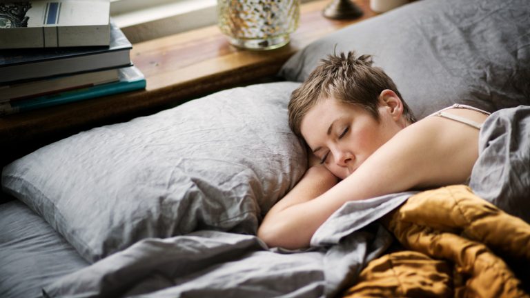 Eine Person liegt in einem Bett mit grauem Bezug und schl&auml;ft, w&auml;hrend eine Hand unter dem Kopf liegt.