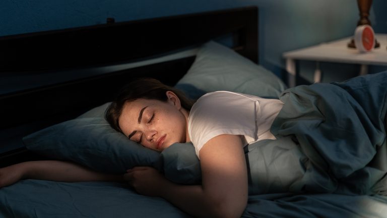 Eine Person liegt in Seitenposition mit geschlossenen Augen im Bett. Ihr Arm liegt unter dem Kissen, auf dem der Kopf ruht.