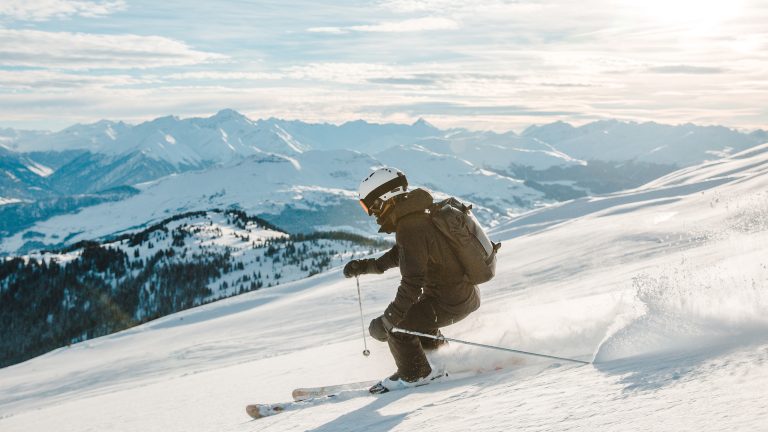 Eine Person f&auml;hrt auf Skiern die Piste herunter. Im Hintergrund sind schneebedeckte Berge zu sehen.