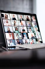 Auf einem Schreibtisch steht ein 2-in-1-Laptop auf dem ein digitales Meeting zu sehen ist. Davor sitzt eine Person, von der nur die Hände zu sehen sind.