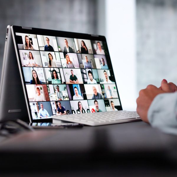 Auf einem Schreibtisch steht ein 2-in-1-Laptop auf dem ein digitales Meeting zu sehen ist. Davor sitzt eine Person, von der nur die H&auml;nde zu sehen sind.