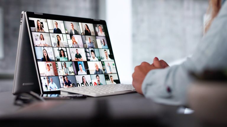 Auf einem Schreibtisch steht ein 2-in-1-Laptop auf dem ein digitales Meeting zu sehen ist. Davor sitzt eine Person, von der nur die Hände zu sehen sind.