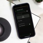 Ein iPhone liegt neben einem Lautsprecher auf einer Tischkante. Darauf sind die AirPlay-Einstellungen mit einer Verbindung zu einem Lautsprecher in einem Wohnzimmer zu sehen. 