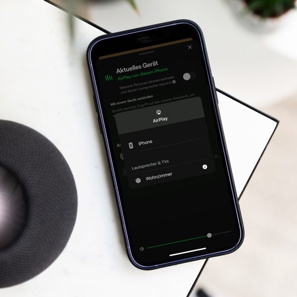 Ein iPhone liegt neben einem Lautsprecher auf einer Tischkante. Darauf sind die AirPlay-Einstellungen mit einer Verbindung zu einem Lautsprecher in einem Wohnzimmer zu sehen.&nbsp;