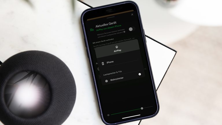 Ein iPhone liegt neben einem Lautsprecher auf einer Tischkante. Darauf sind die AirPlay-Einstellungen mit einer Verbindung zu einem Lautsprecher in einem Wohnzimmer zu sehen. 
