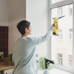 Eine Person steht in einer K&uuml;che vor dem Fenster und zieht mit einem Fenstersauger die Fensterscheibe von innen ab.