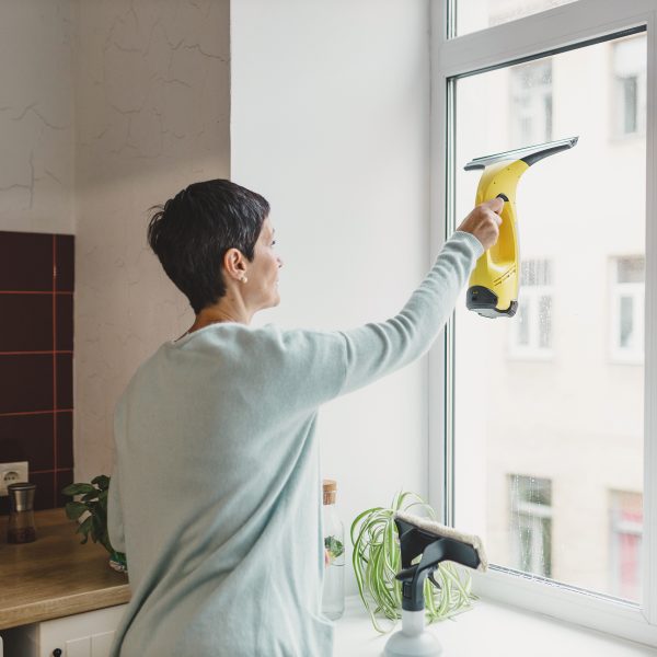 Eine Person steht in einer K&uuml;che vor dem Fenster und zieht mit einem Fenstersauger die Fensterscheibe von innen ab.