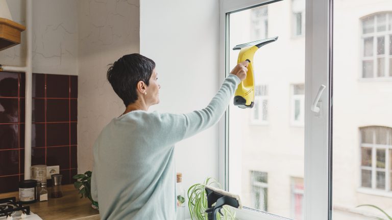 Eine Person steht in einer K&uuml;che vor dem Fenster und zieht mit einem Fenstersauger die Fensterscheibe von innen ab.