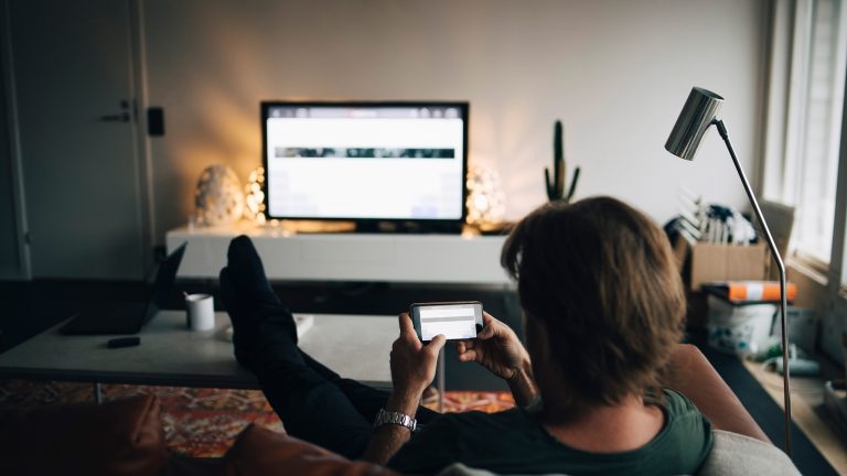 Eine Person sitzt auf einer Couch in einem Wohnzimmer und h&auml;lt ihr Smartphone quer in den H&auml;nden. Auf einem Fernseher vor ihr sieht man den gleichen Inhalt, wie auf dem Smartphone.