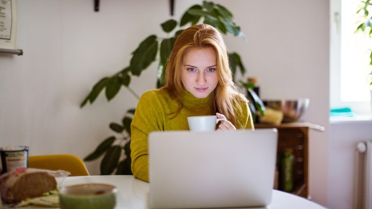 Eine Person sitzt am Küchentisch vor ihrem Laptop. In der linken Hand hält die Person eine Tasse.