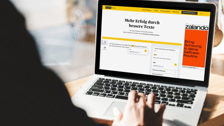 Ein Notebook steht auf einer hölzernen Schreibtischplatte. Im Browser ist der Duden-Mentor geöffnet.