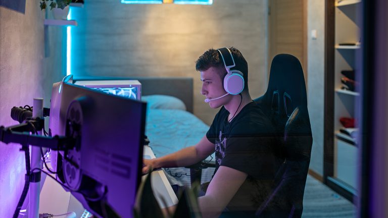 Eine Person mit einem weißen Headset auf dem Kopf sitzt vor einem großen Gaming-PC-Setup, das bunt beleuchtet ist.