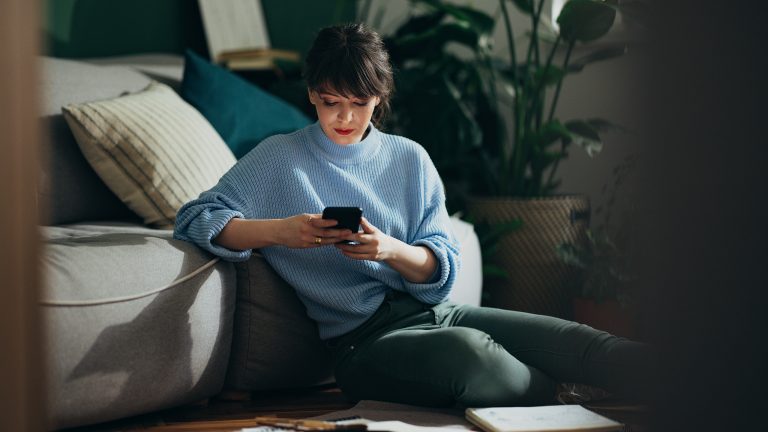 Eine Person sitzt im Halbdunkel auf dem Boden vor dem Sofa und h&auml;lt ein Smartphone in den H&auml;nden.