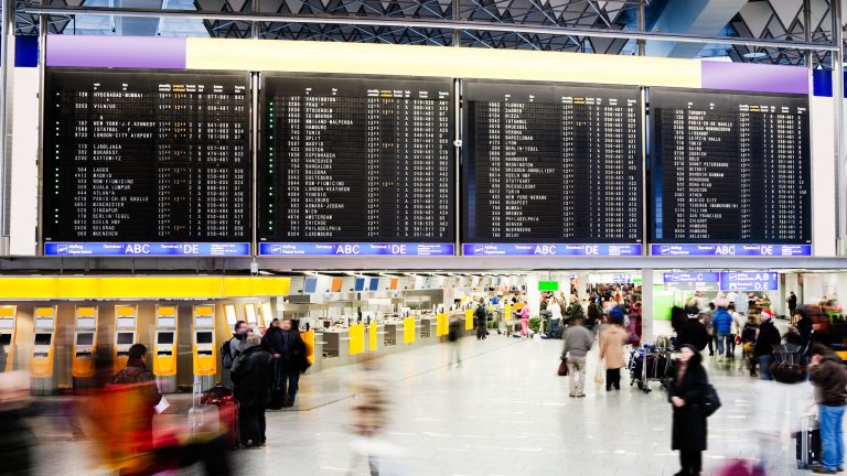 Eine gut besuchte Flughafenhalle über der eine Fluganzeigentafel hängt.