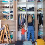 Eine Person steht mit zwei Kindern vor einem deckenhohen Kleiderschrank mit vielen F&auml;chern und ge&ouml;ffneten T&uuml;ren.