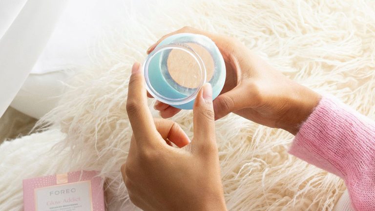 Die H&auml;nde einer Person &ouml;ffnen das FOREO UFO &uuml;ber einer wei&szlig;en Kuscheldecke. Daneben liegt die Verpackung der Maske &bdquo;Glow Addict&ldquo; von FOREO.