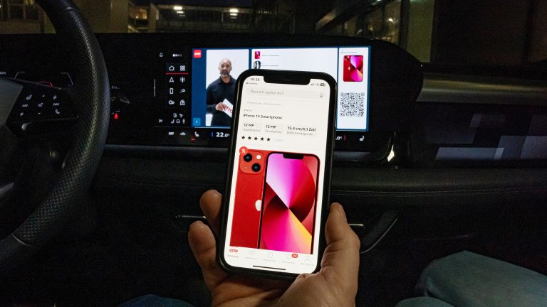 Eine Person sitzt in einem Audi Q6 e-tron und h&auml;lt ein Smartphone in der Hand. Darauf ist eine Produktdetailseite aus dem OTTO-Shop zusehen und im Hintergrund l&auml;uft auf dem integrierten Display ein Live-Shopping-Stream.