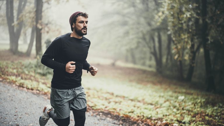 Ein Mensch joggt in Sportklamotten auf einer Stra&szlig;e in einem diesigen Wald.