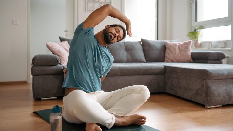 Eine Person sitzt im Schneidersitz vor einem Sofa auf einer Yogamatte und dehnt sich.