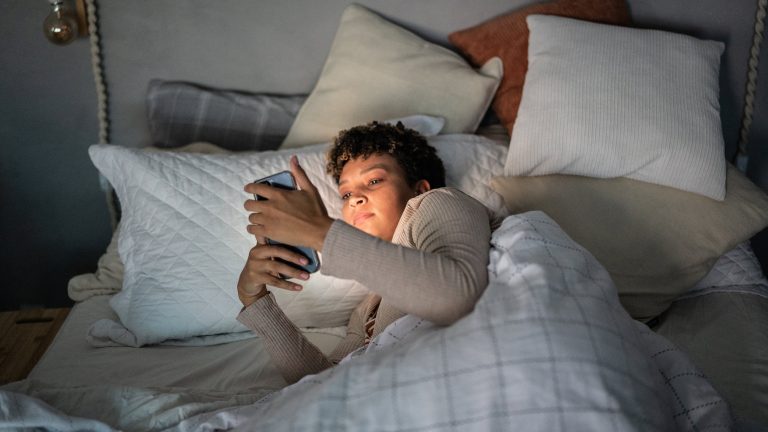 Eine Person liegt in einem Bett mit vielen Kissen, h&auml;lt ihr Smartphone mit beiden H&auml;nden vors Gesicht und wird davon erleuchtet.