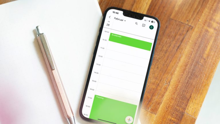 Ein Smartphone liegt halb auf einem Notizbuch, halb auf einem Holztisch. Darauf ist eine Tages&uuml;bersicht eines Kalenders mit zwei Terminen darin zu sehen. Neben dem Handy liegt ein Stift.