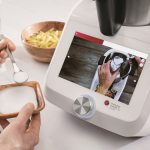 Die H&auml;nde einer Person halten eine kleine Sch&uuml;ssel und einen kleinen L&ouml;ffel, der in die Monsieur Cuisine smart-K&uuml;chenmaschine gegeben werden soll, die auf der K&uuml;chentheke steht.