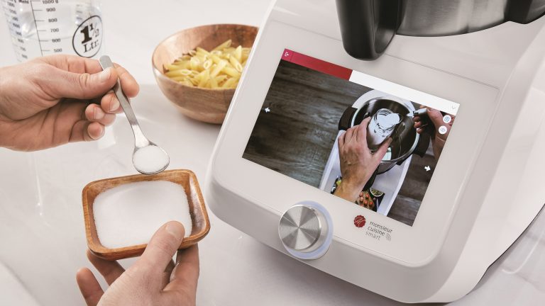 Die Hände einer Person halten eine kleine Schüssel und einen kleinen Löffel, der in die Monsieur Cuisine smart-Küchenmaschine gegeben werden soll, die auf der Küchentheke steht.