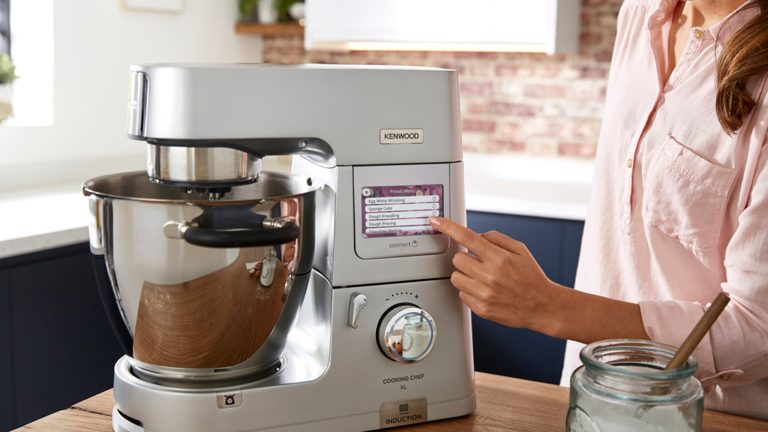 Eine Person bedient per LED-Bedienfeld eine silberne Küchenmaschine von Kenwood, die auf einer Küchentheke aus Holz steht.