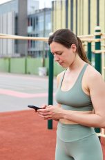 Eine Person im Trainings-Outfit steht auf einer Calisthenics-Anlage in einem Park. Sie schaut auf ihr Smartphone, das sie in beiden H&auml;nden h&auml;lt.