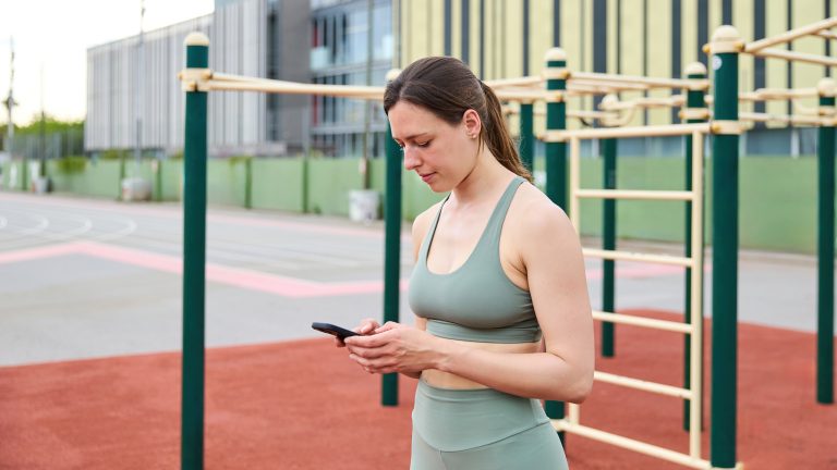 Eine Person im Trainings-Outfit steht auf einer Calisthenics-Anlage in einem Park. Sie schaut auf ihr Smartphone, das sie in beiden Händen hält.