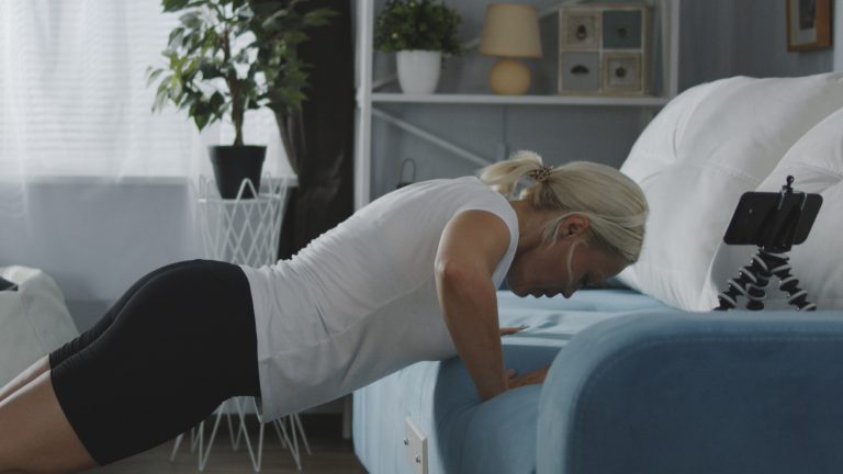 Eine Person macht Push-Ups. Das Sofa dient dabei als Stütze. Das Smartphone steht auf einem Ständer auf der Couch.